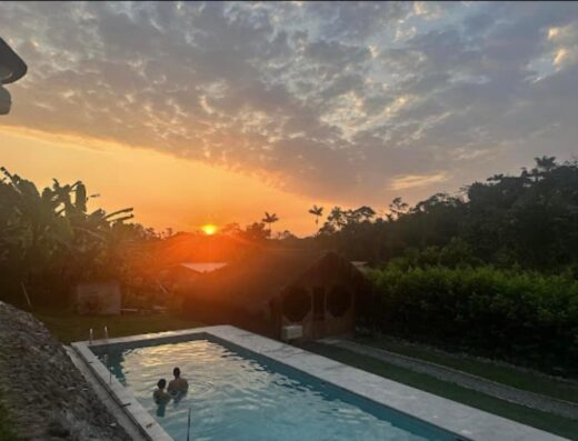 A gorgeous orange sunset is on the horizon with palm trees on the skyline, and their images reflected off of a glistening pool at an ayahuasca retreat in the jungle
